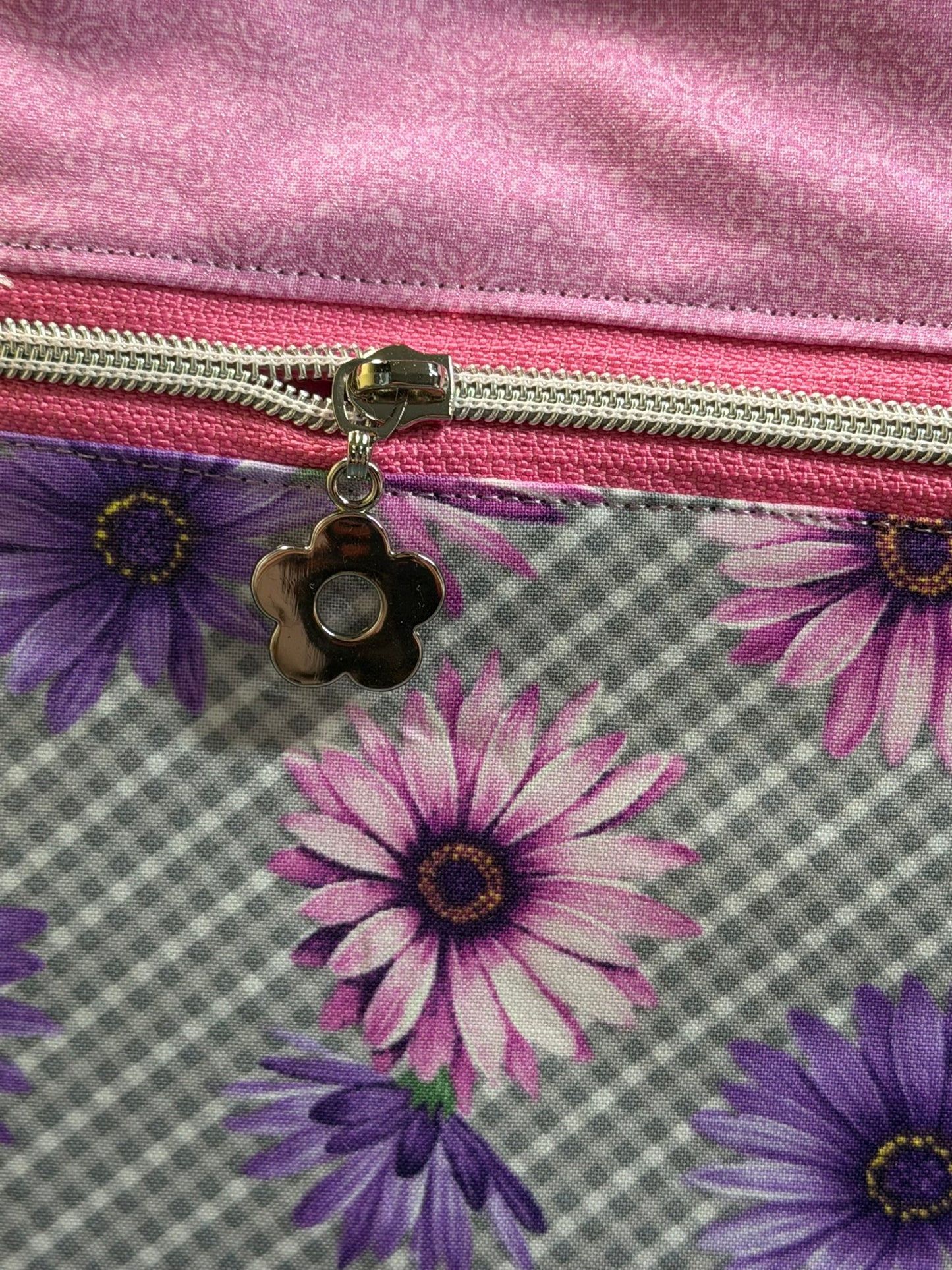 Close-up of a fabric zippered pouch or bag with a pink satin-like top section, a pink zipper track, a silver zipper pull, and a floral lower section featuring purple and pink daisy-like flowers on a gray plaid background.