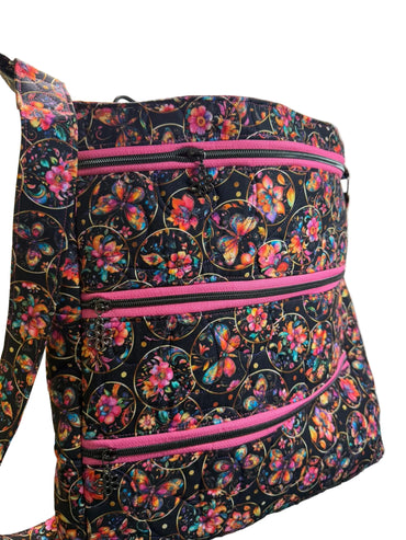 A fabric tote bag with a black background and a colorful butterfly and floral pattern, featuring three horizontal pink zippers.