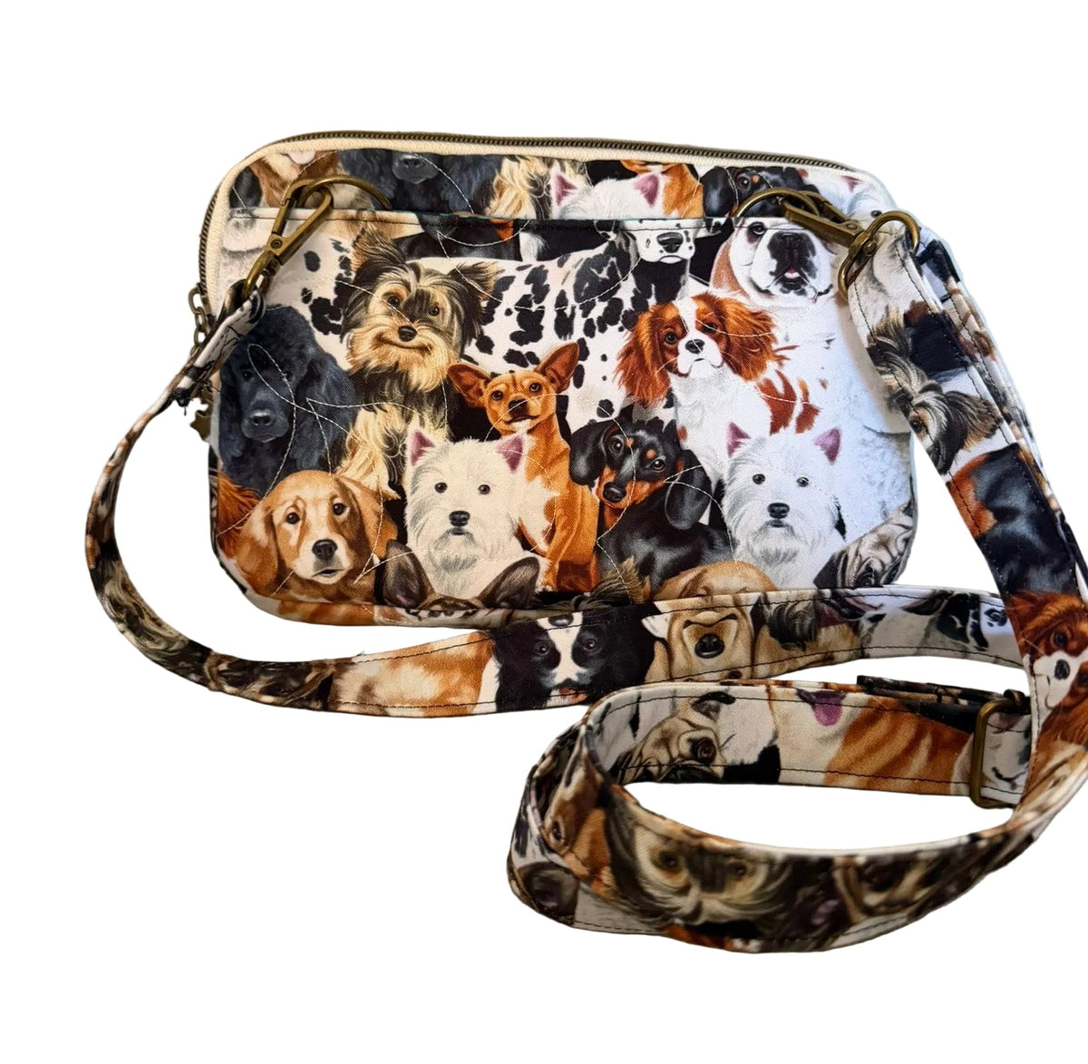 A small crossbody handbag with a colorful print of various dog faces, featuring a matching patterned strap and gold-tone hardware, set against a textured off-white wall and gray floor.