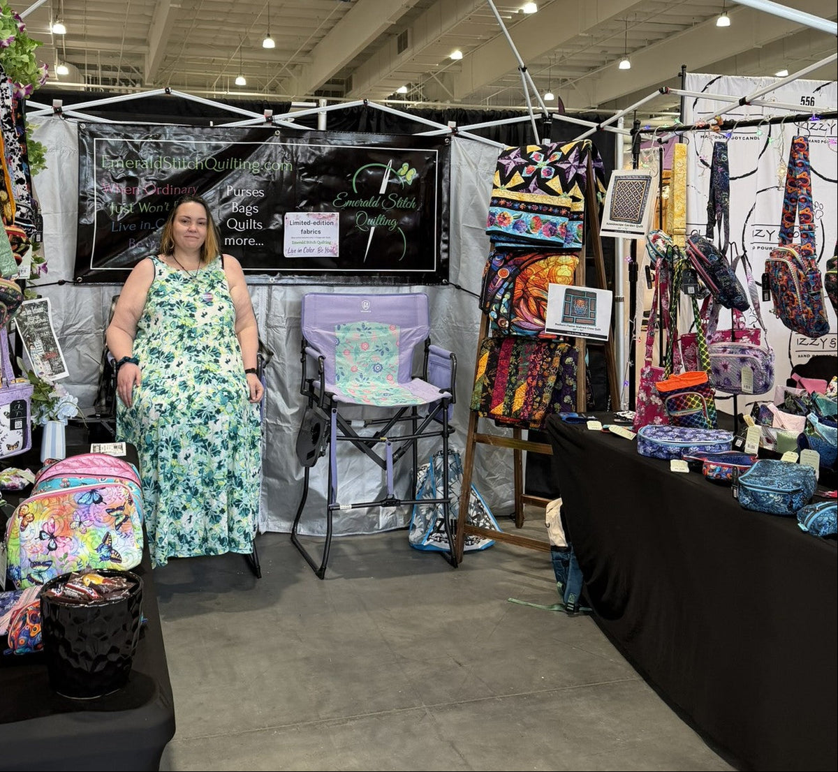 A woman stands at a craft fair booth surrounded by colorful handmade bags, backpacks, and textile items. She is wearing a green and white floral dress, with a black display backdrop behind her and a table covered in patterned goods to her right.