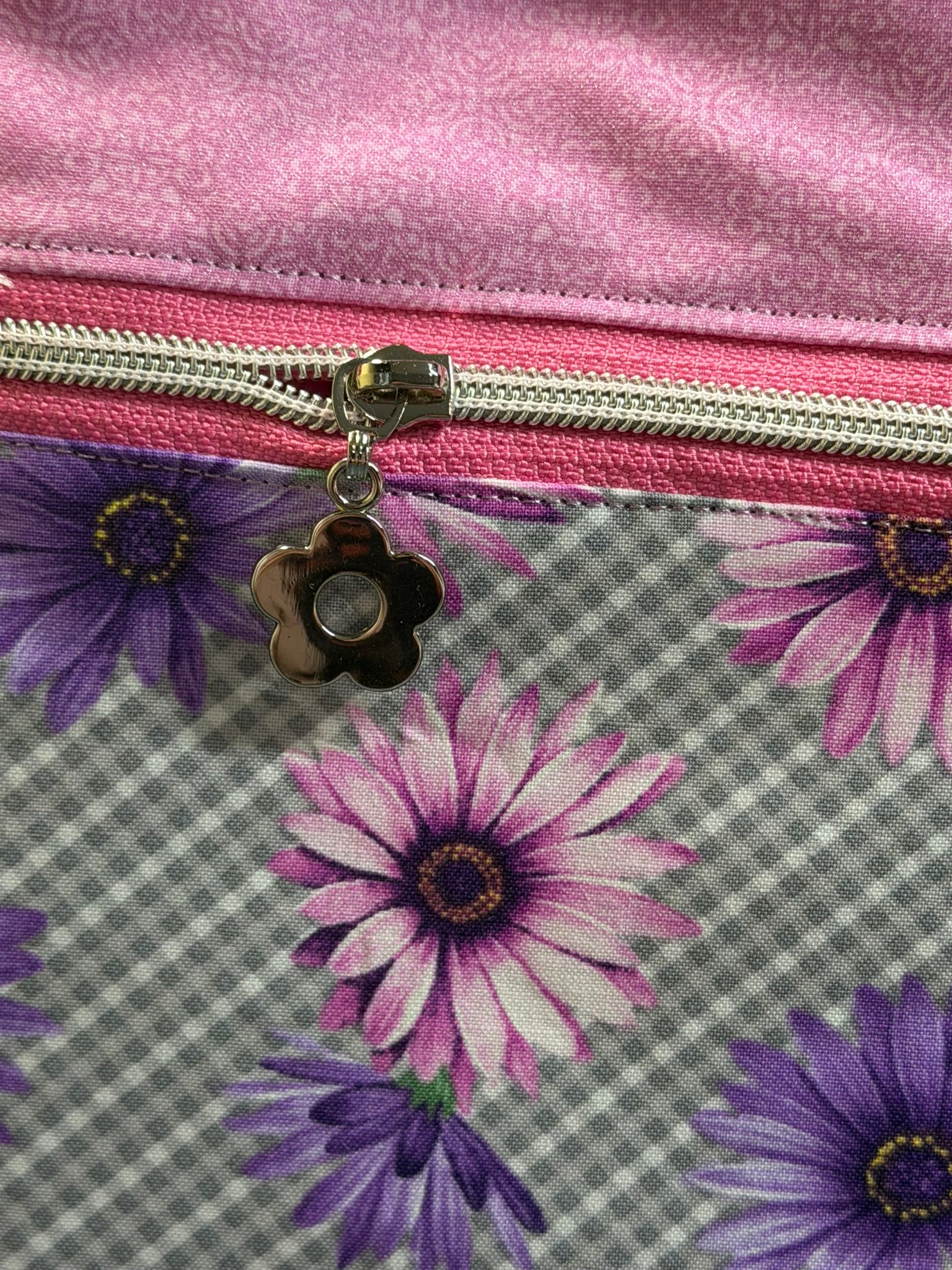 Close-up of a fabric zippered pouch or bag with a pink satin-like top section, a pink zipper track, a silver zipper pull, and a floral lower section featuring purple and pink daisy-like flowers on a gray plaid background.