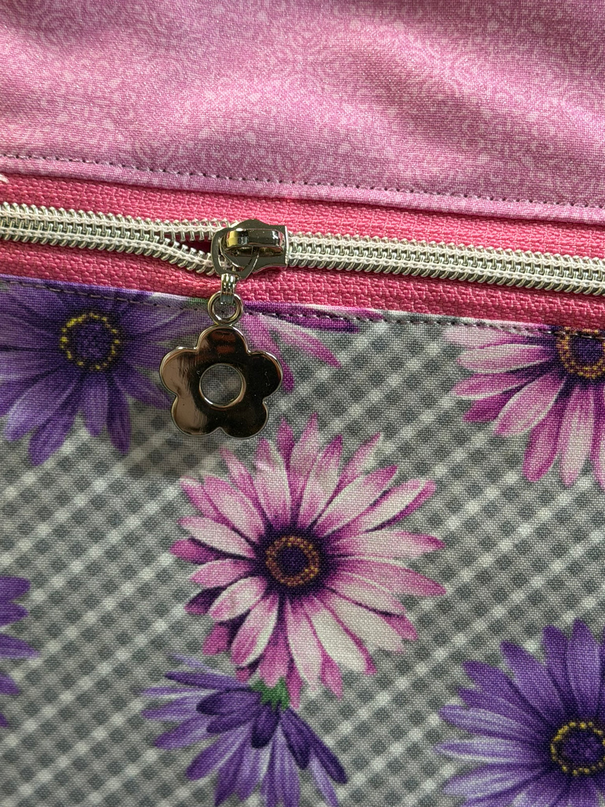 Close-up of a fabric zippered pouch or bag with a pink satin-like top section, a pink zipper track, a silver zipper pull, and a floral lower section featuring purple and pink daisy-like flowers on a gray plaid background.