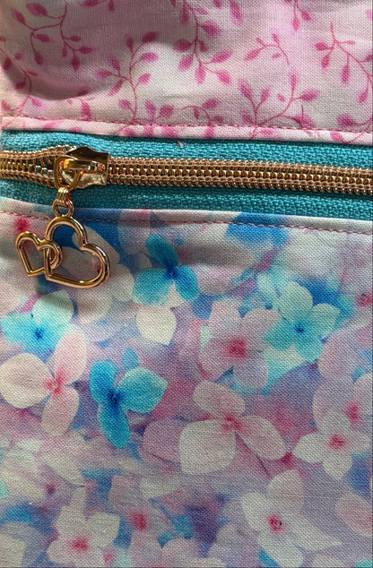 Close-up of a fabric pouch or bag with a floral print in pink, blue, and white, featuring a gold zipper and a gold heart-shaped zipper pull.