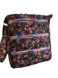 A fabric tote bag with a black background and a colorful floral and butterfly print, featuring three horizontal iridescent zippered pockets.