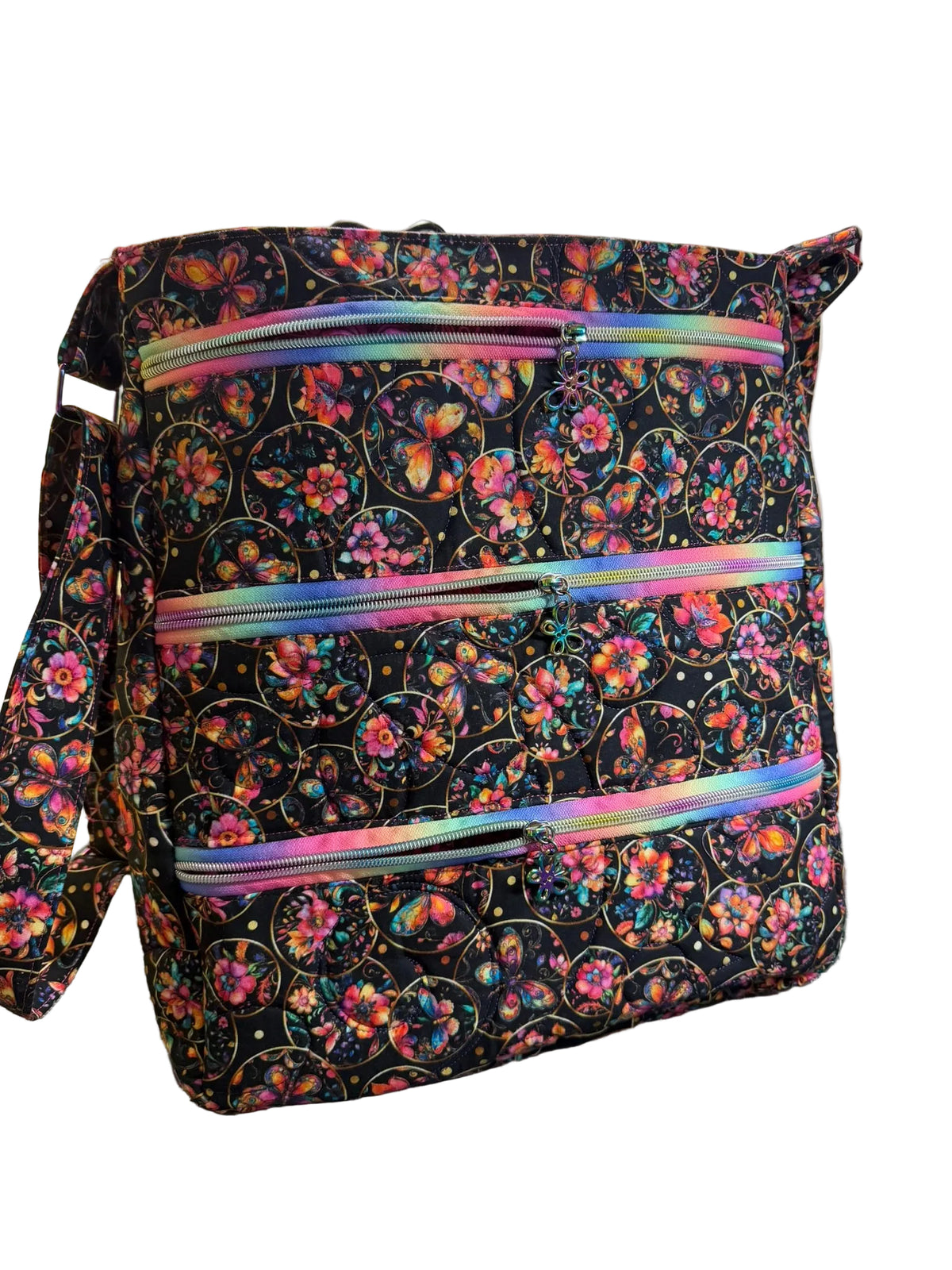 A fabric tote bag with a black background and a colorful floral and butterfly print, featuring three horizontal iridescent zippered pockets.