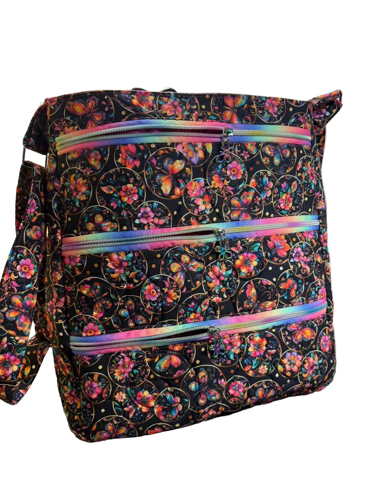 A fabric tote bag with a black background and a colorful floral and butterfly print, featuring three horizontal iridescent zippered pockets.