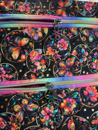 Close-up of a fabric bag or tote with a black background covered in colorful butterfly and floral motifs, plus two rainbow-colored zippers running horizontally.