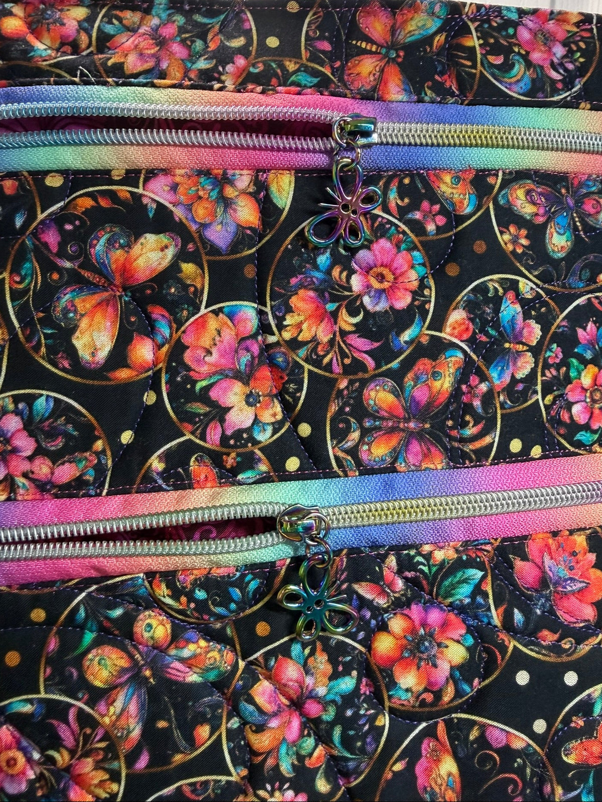 Close-up of a fabric bag or tote with a black background covered in colorful butterfly and floral motifs, plus two rainbow-colored zippers running horizontally.