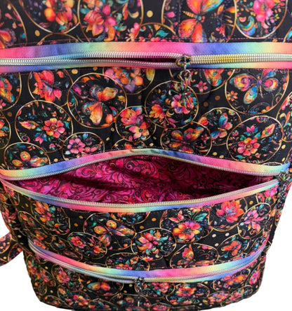 A close-up of a fabric zippered pouch or organizer with a black background, colorful butterfly and floral print, and iridescent rainbow zipper trim.