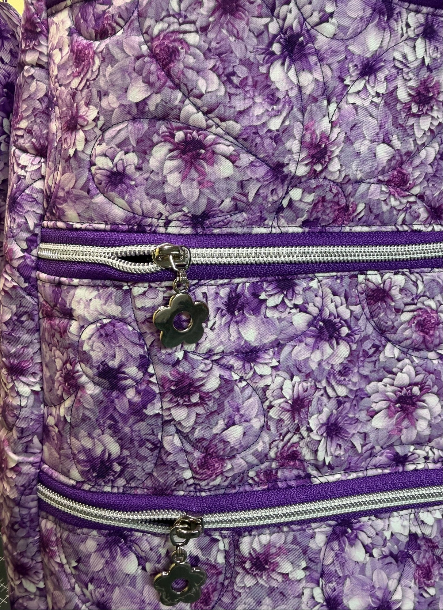 Close-up of a quilted messenger bag with a purple and white floral pattern, featuring two horizontal zippers with silver metal teeth and dark purple flower-shaped zipper pulls.