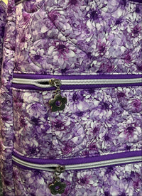 Close-up of a quilted messenger bag with a purple and white floral pattern, featuring two horizontal zippers with silver metal teeth and dark purple flower-shaped zipper pulls.