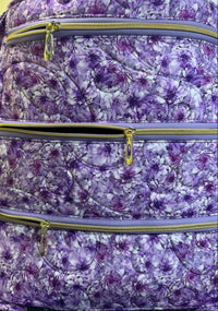 A close-up of a quilted bag featuring a purple and white floral pattern, with three horizontal zippered compartments and gold-tone zipper pulls.