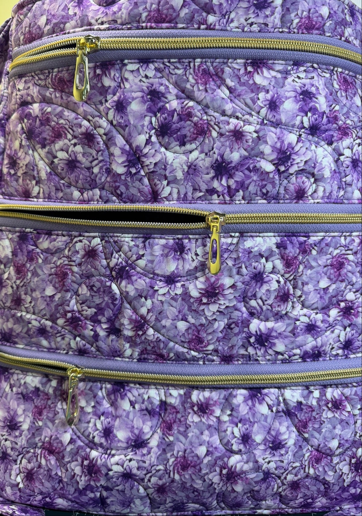 A close-up of a quilted bag featuring a purple and white floral pattern, with three horizontal zippered compartments and gold-tone zipper pulls.