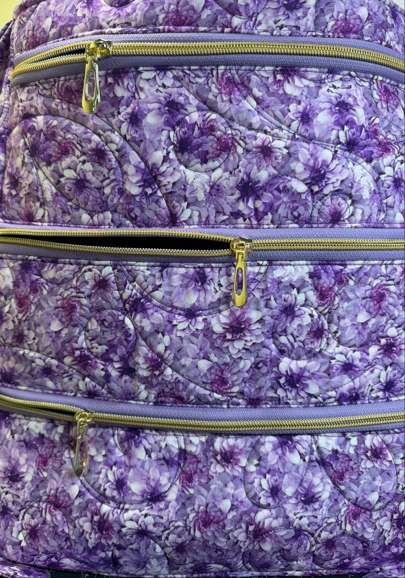 A close-up of a quilted bag featuring a purple and white floral pattern, with three horizontal zippered compartments and gold-tone zipper pulls.