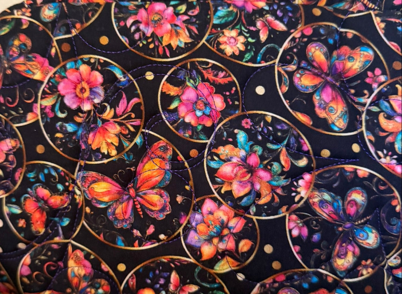 A fabric zippered pouch with a black background and a colorful print of butterflies and flowers inside gold circular frames.